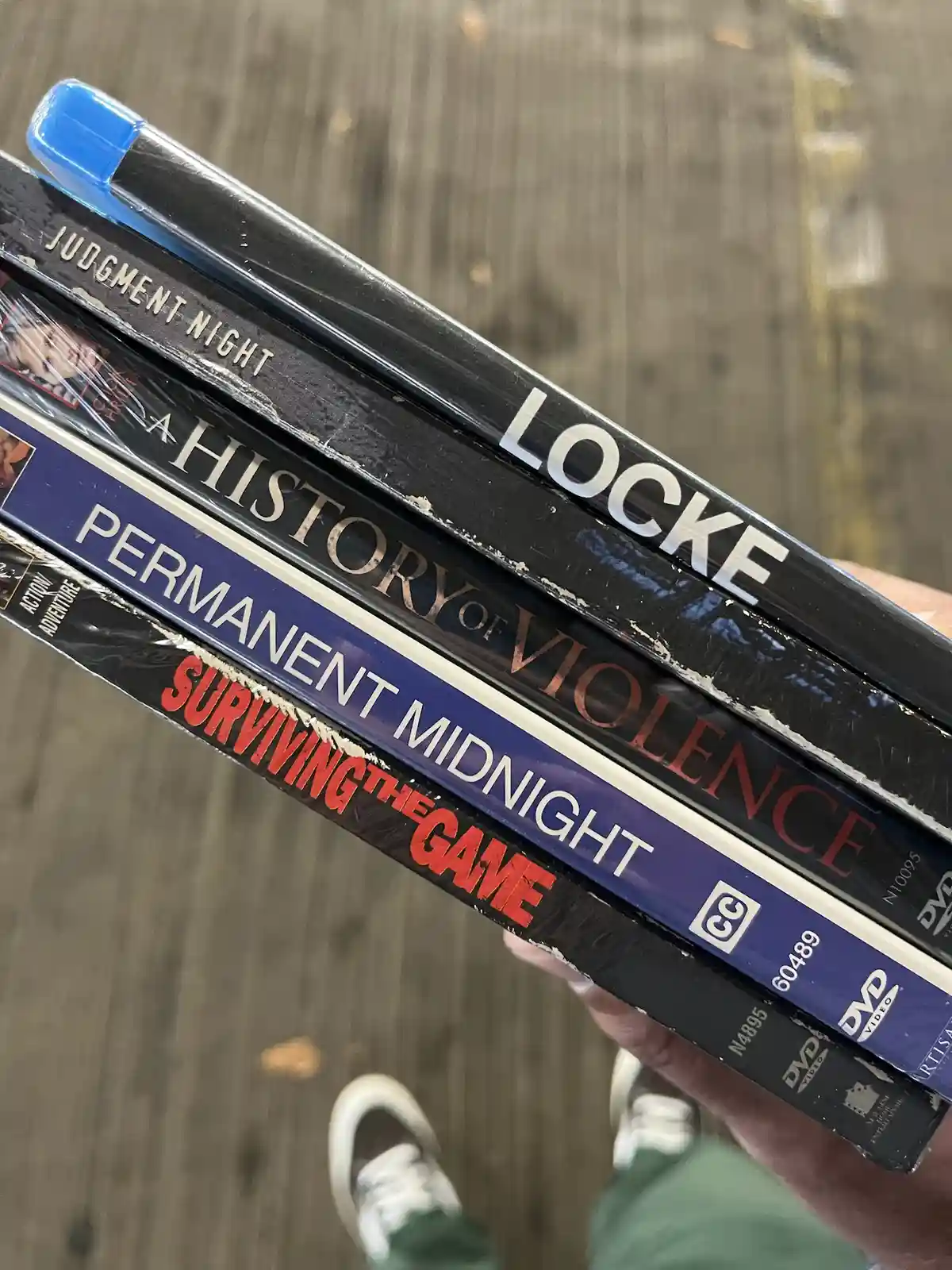 A hand holds a stack of five worn, scratched DVD cases with titles like 'Judgment Night,' 'Locke,' 'A History of Violence,'…