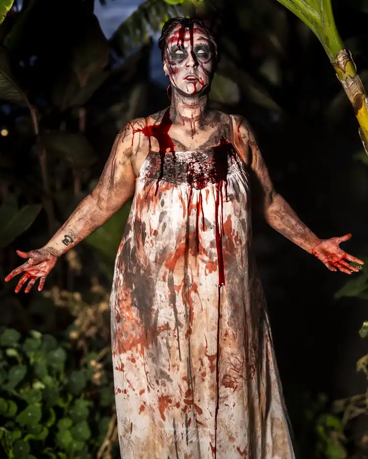 A person in a white dress covered in fake blood stands with arms outstretched in a dark, leafy outdoor setting. Their face…