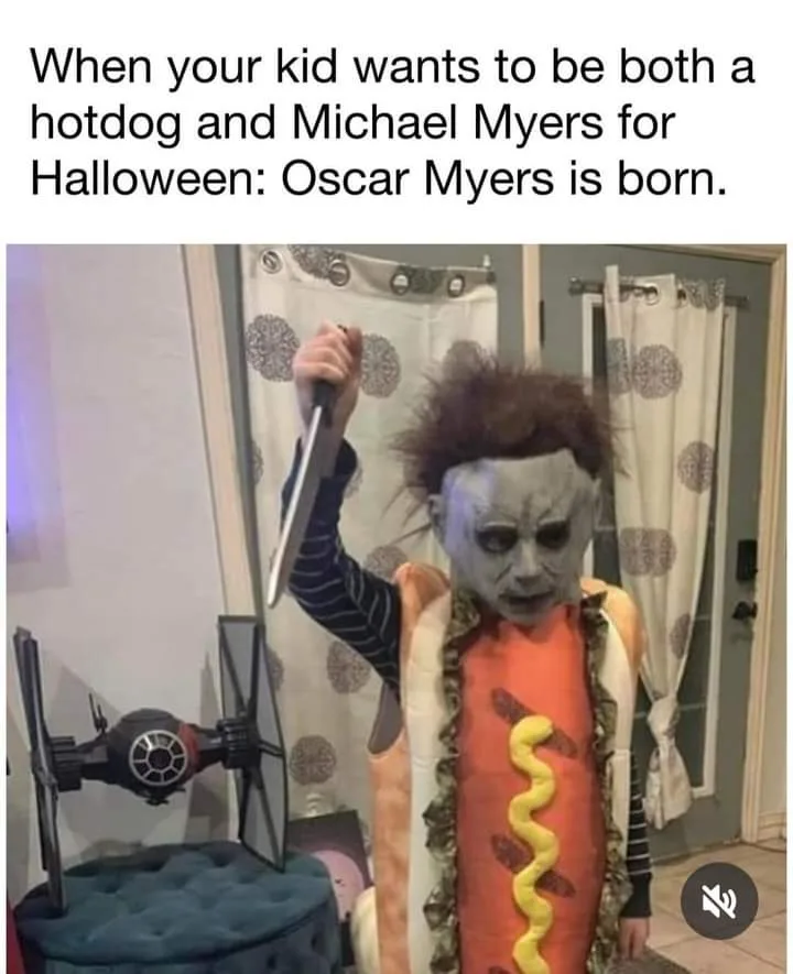 A child dressed as a hot dog with mustard, wearing a Michael Myers mask and holding a large knife, standing in a room with a…
