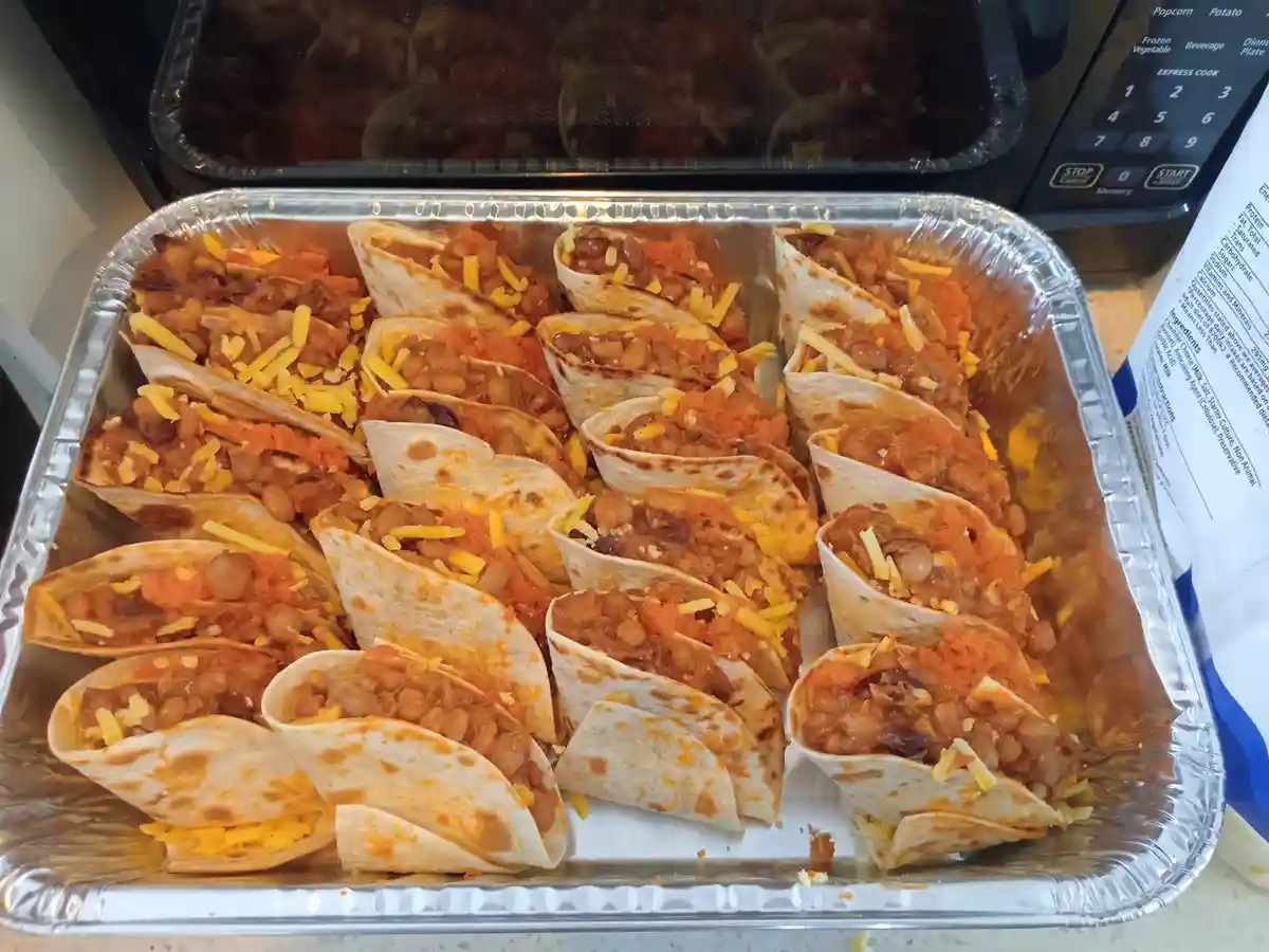 A large aluminum tray holds numerous small, folded tacos filled with beans, cheese, and sauce, arranged neatly. In the…