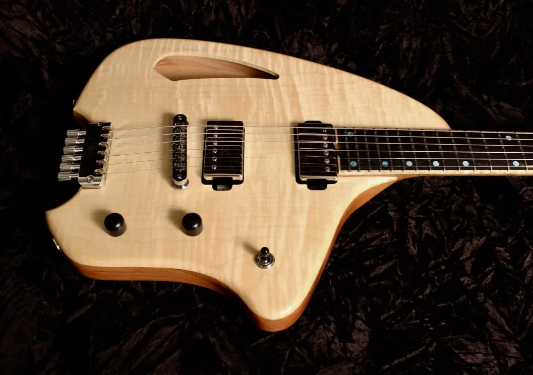 a light-colored electric guitar with a unique, angular body shape resembling a spaceship, resting on a dark, textured fabric…