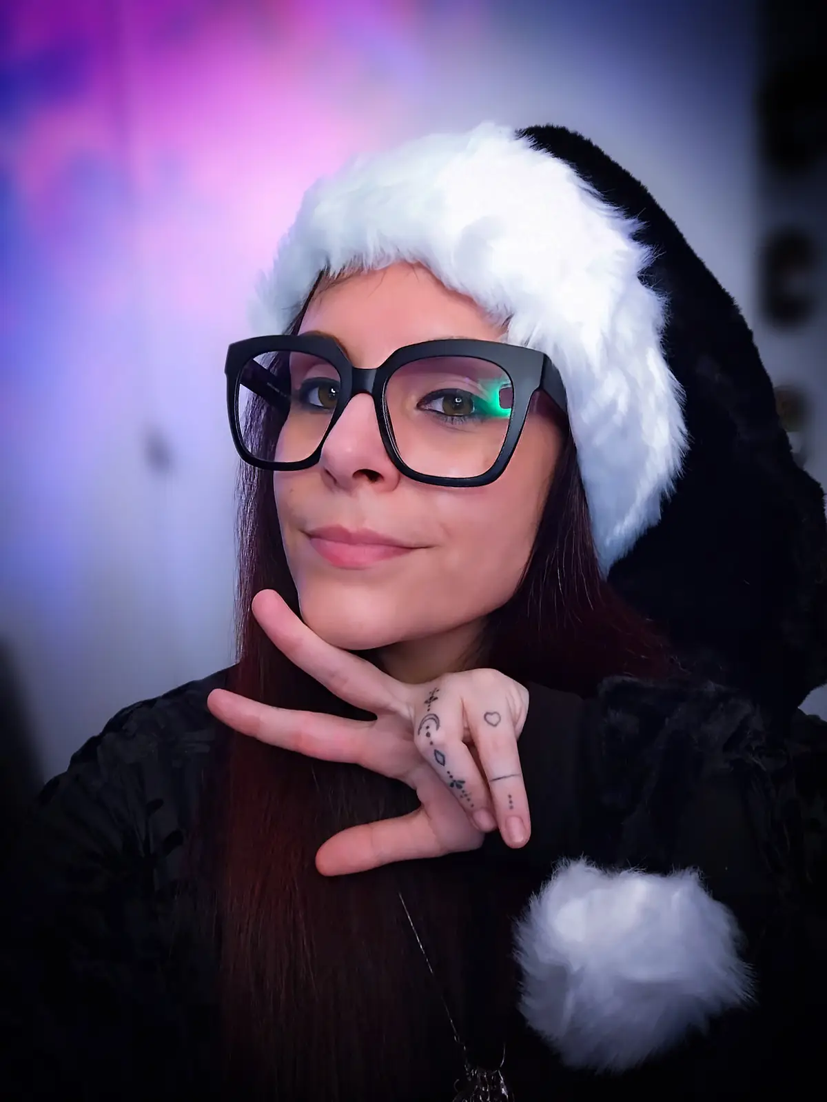 A person with dark hair and large black glasses wears a black and white Santa hat and a black outfit with a white pom-pom…