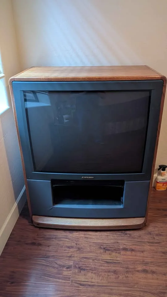 A vintage Mitsubishi CRT television with a wooden frame sits on a hardwood floor, its screen off and reflecting the room…
