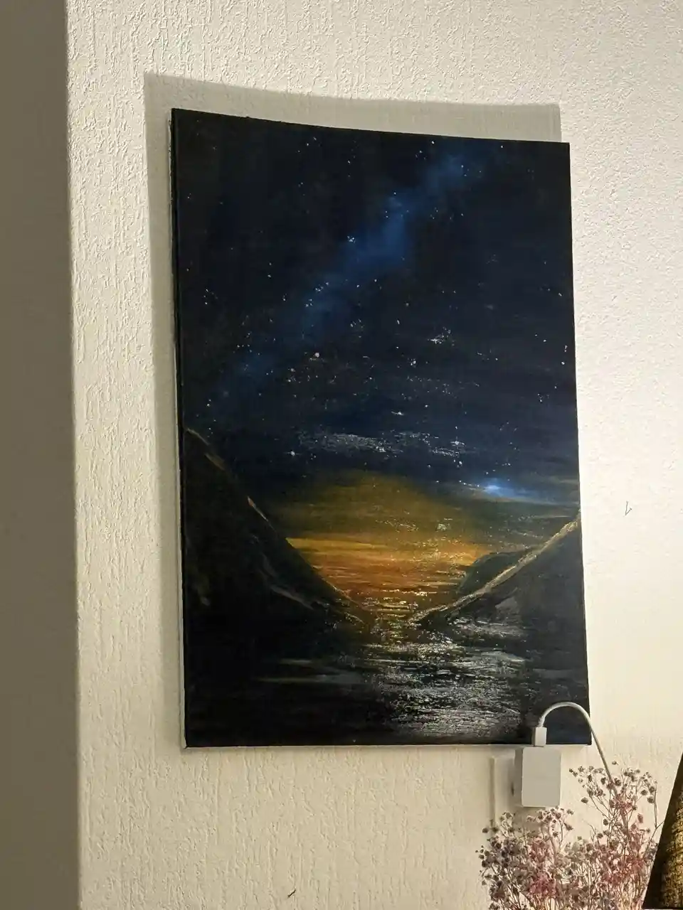 A painting of a starry night sky over a valley hangs on a white wall, with a white USB cable plugged into an outlet below it…
