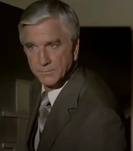 a still image of a man with gray hair wearing a suit and tie, looking directly at the camera with a serious, slightly…