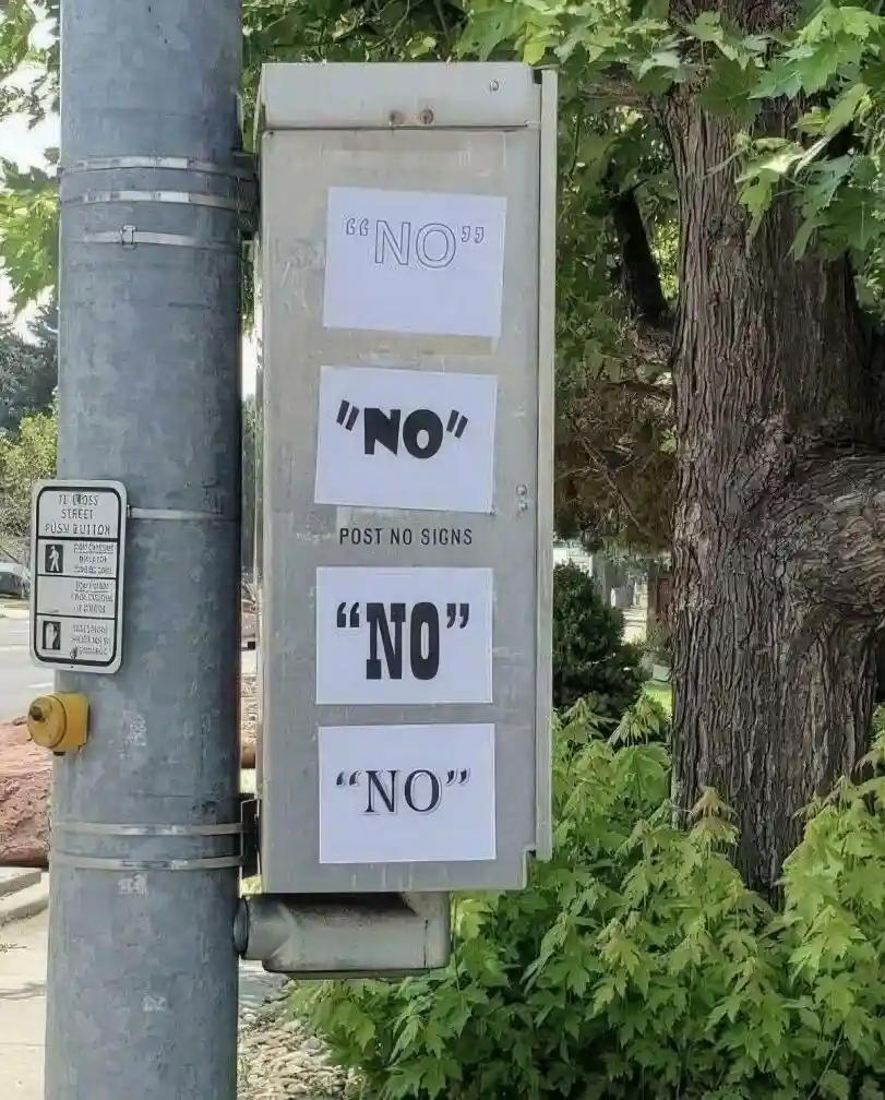 A metal box attached to a pole has four white paper slips taped vertically, each displaying the word 'NO' in different fonts…