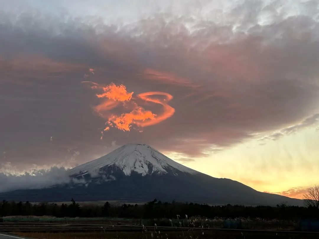 a photograph of mount fuji at sunset, its snow-capped peak silhouetted against a dramatic sky with clouds glowing orange. a…