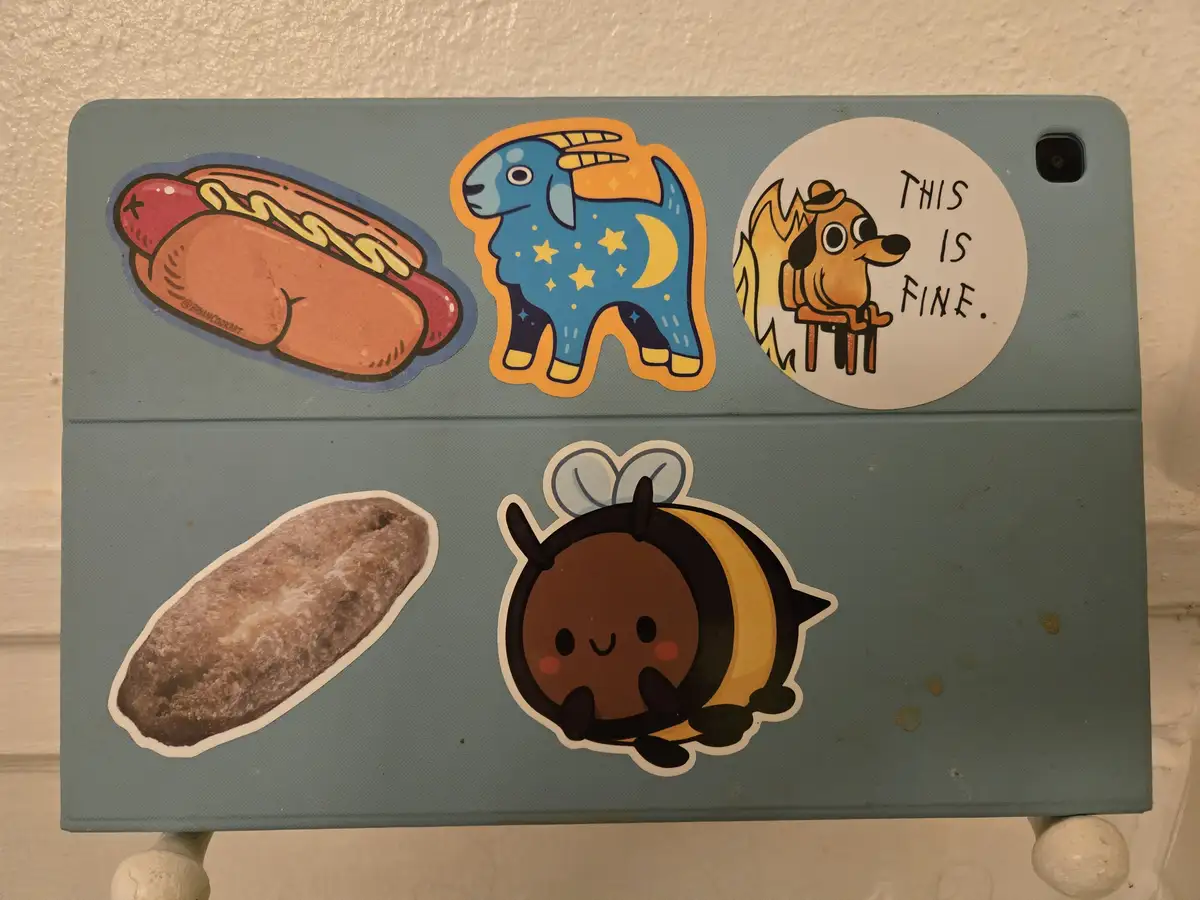 A light blue tablet case is decorated with five stickers: a hot dog with mustard, a blue goat with stars and a crescent…