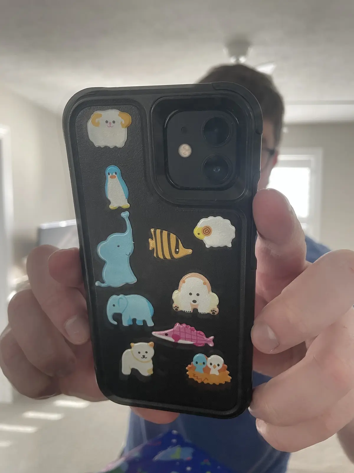 A person holds up a black phone case covered in colorful, cute animal stickers including sheep, penguins, elephants, fish…