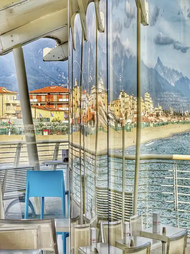 The image shows a real beachfront cafe with tables, chairs, and a view of the sea and distant mountains, but the large glass…