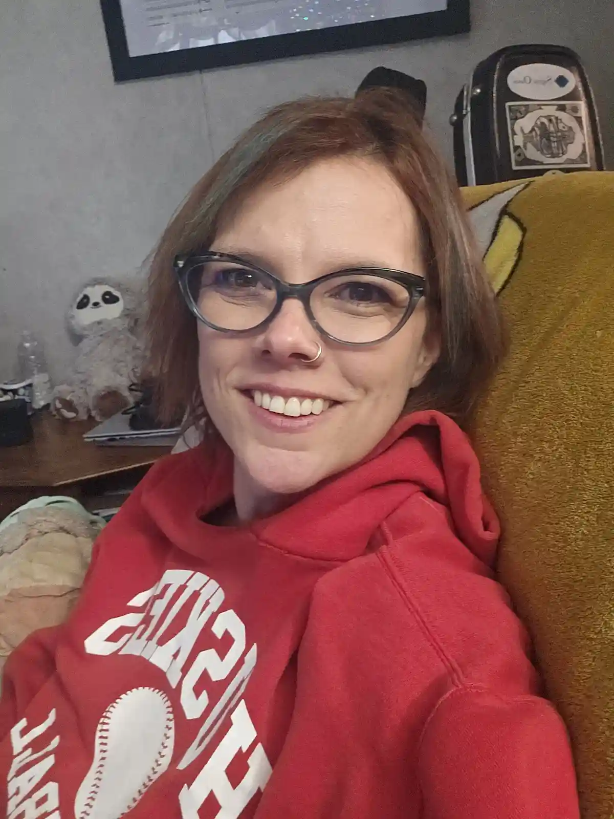 A person with glasses and a nose ring smiles while wearing a red hoodie with a baseball graphic, seated on a yellow couch…