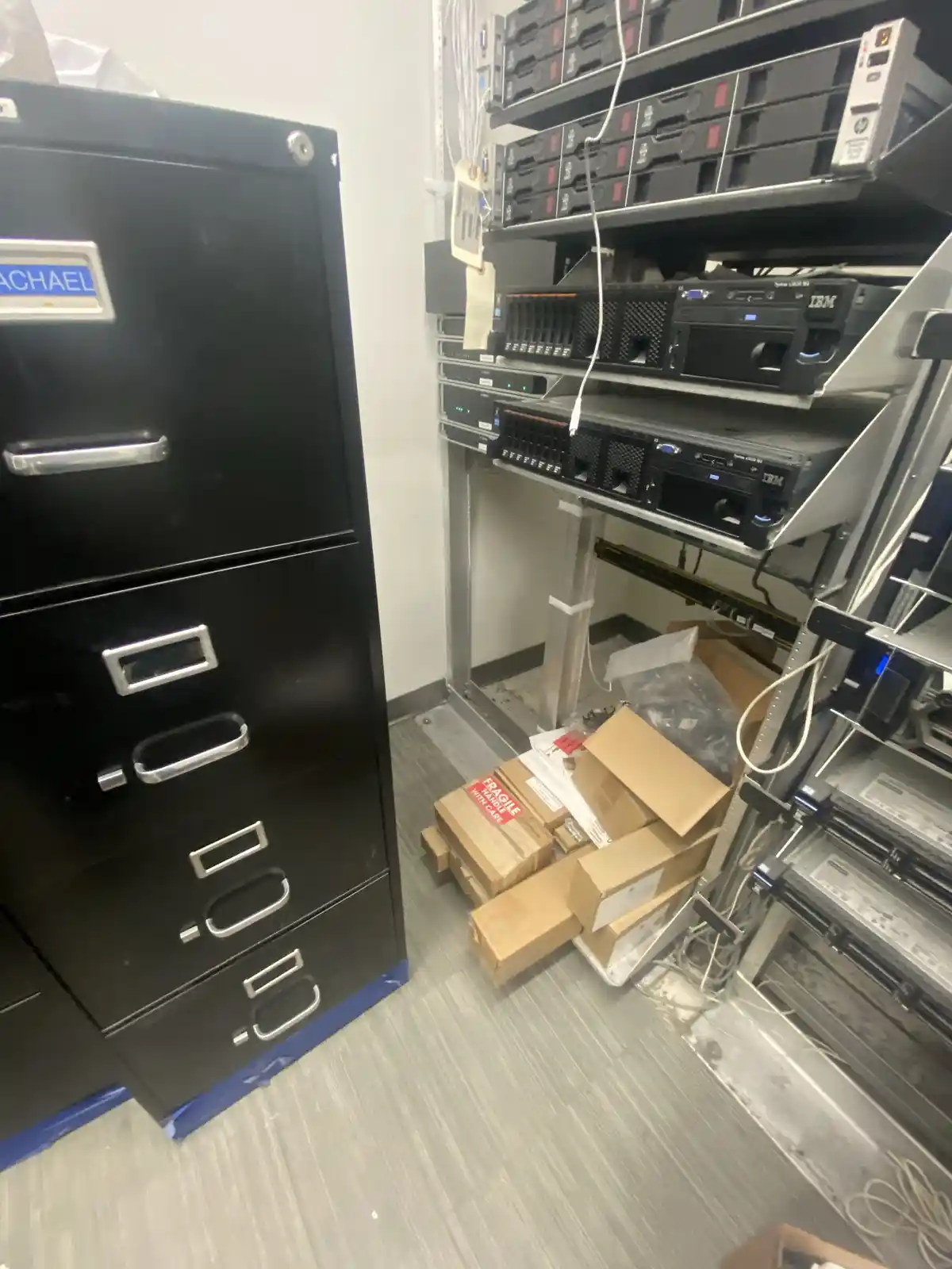 A black filing cabinet with a nameplate reading 'MICHAEL' stands next to a server rack filled with IBM equipment and…