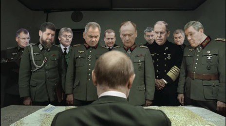 A group of stern-looking military officers in green uniforms stand around a table with a map, all looking down at it. The…