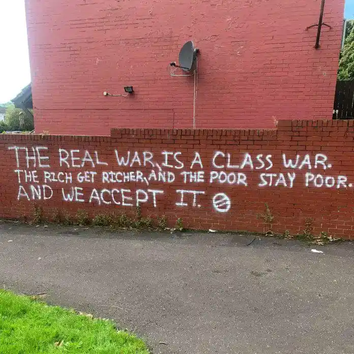 A red brick wall with white spray-painted text stating 'THE REAL WAR IS A CLASS WAR. THE RICH GET RICHER AND THE POOR STAY…