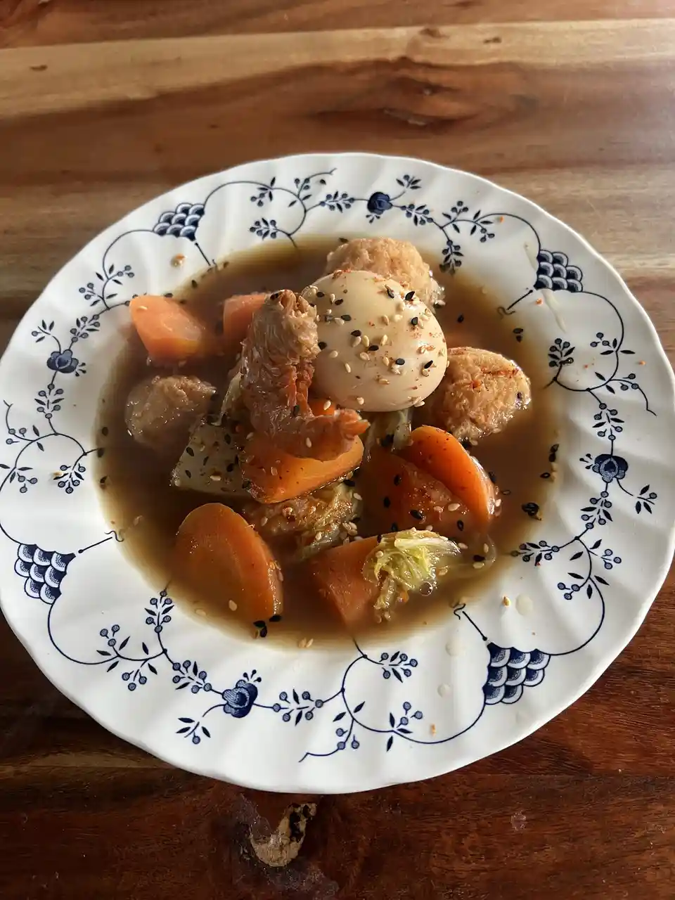 a close-up photo shows a white ceramic bowl with blue floral patterns, filled with a brown broth containing sliced carrots…