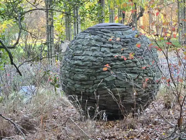 A large, spherical stone structure resembling a dome or igloo sits in an autumnal garden, surrounded by trees with red and…
