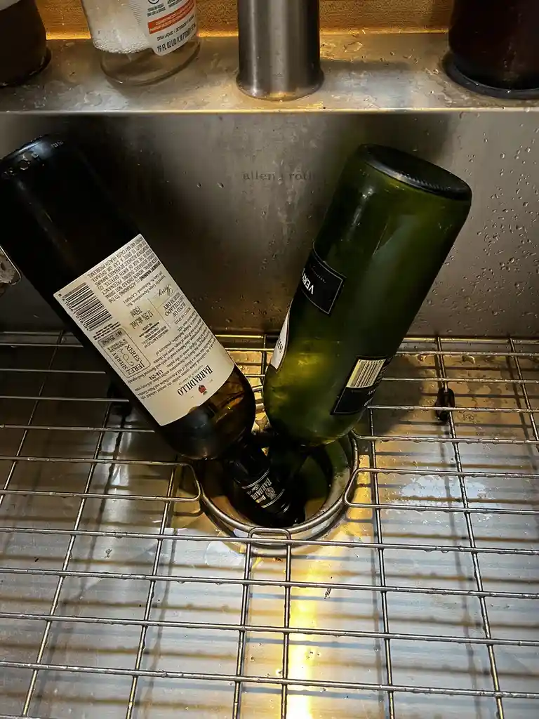 Two empty wine bottles are stuck together in a kitchen sink drain, with water droplets visible on the stainless steel…