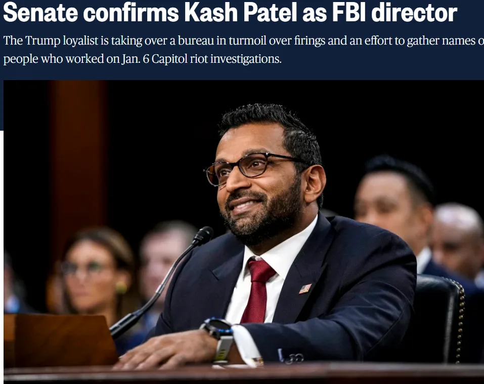 A man in a dark suit, red tie, and glasses smiles while seated at a wooden table with a microphone in front of him. The…