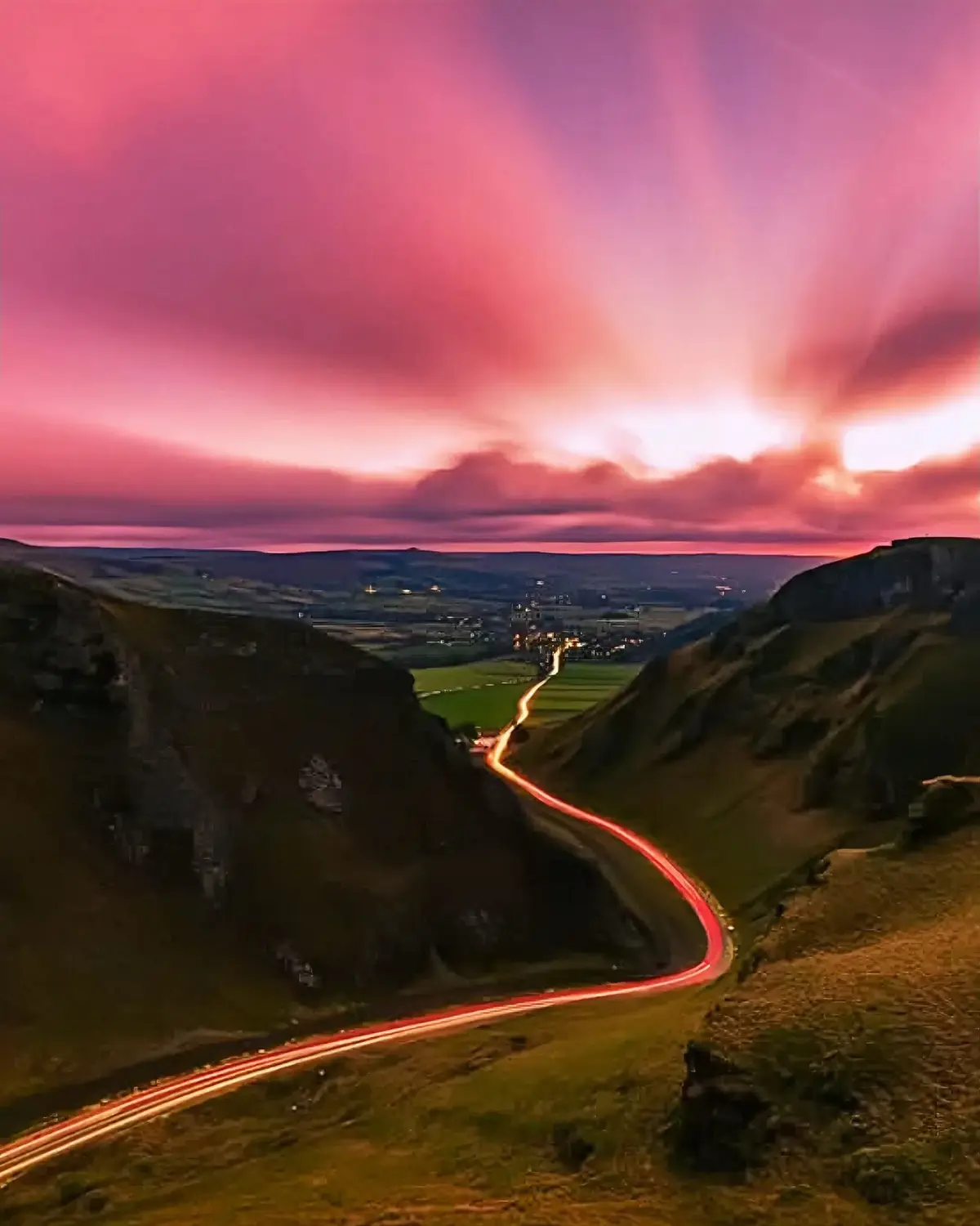 A long-exposure photograph captures a winding road through a valley at dusk, with streaks of red taillights from moving cars…