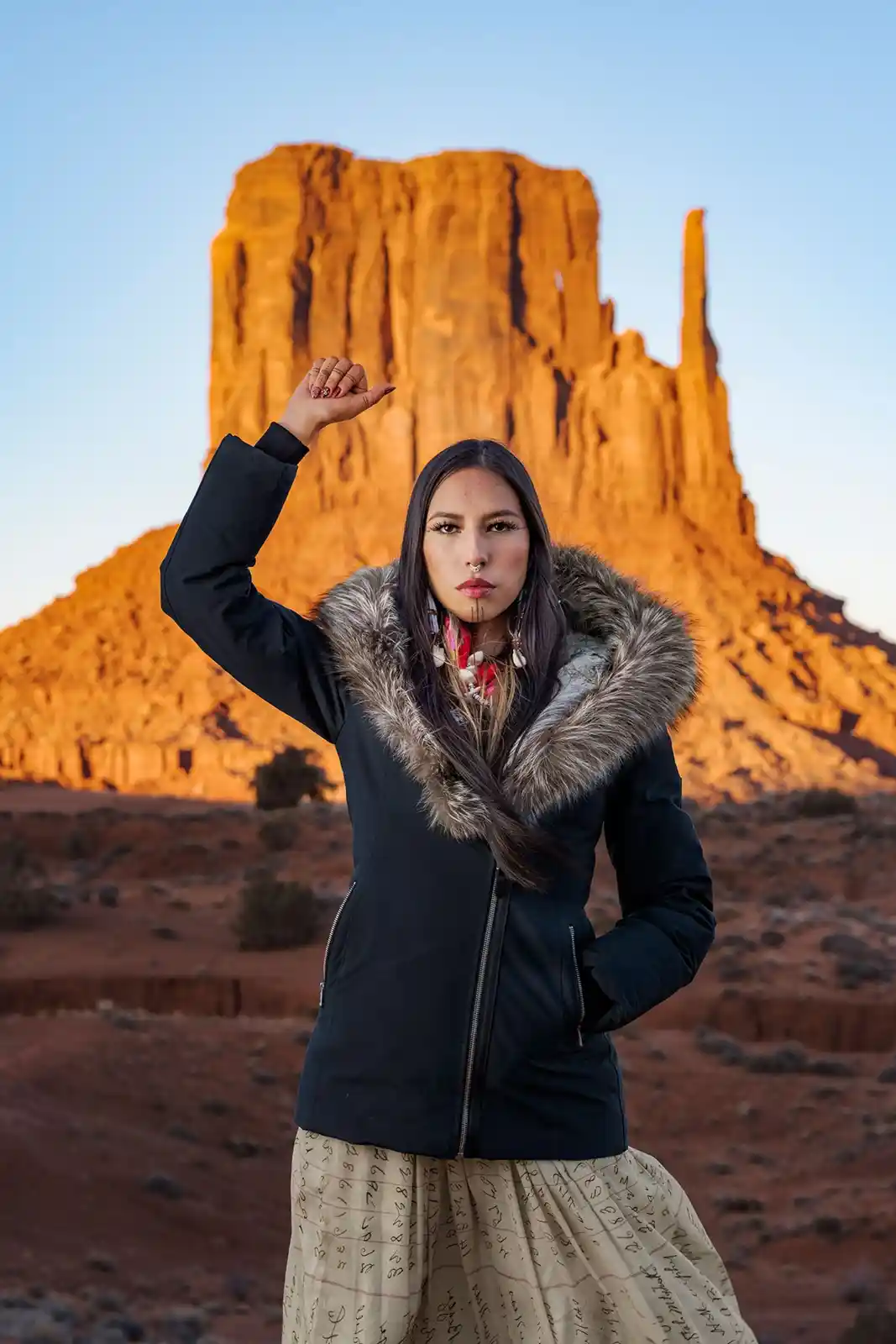 A woman stands in a desert landscape with large orange rock formations behind her, wearing a black jacket with a fur-lined…