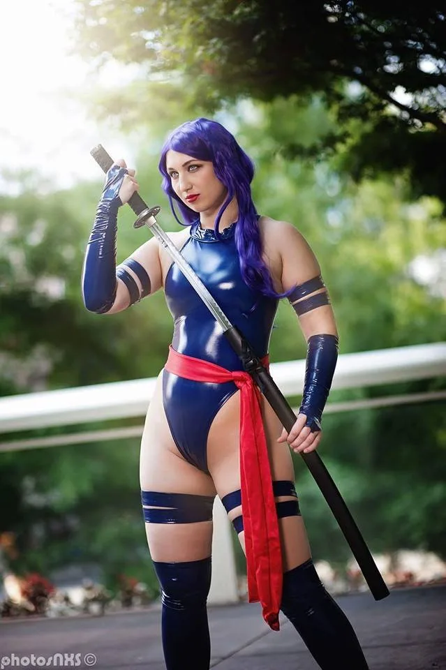 A person with purple hair and blue latex outfit holds a katana outdoors, wearing black arm and leg bands and a red sash. The…