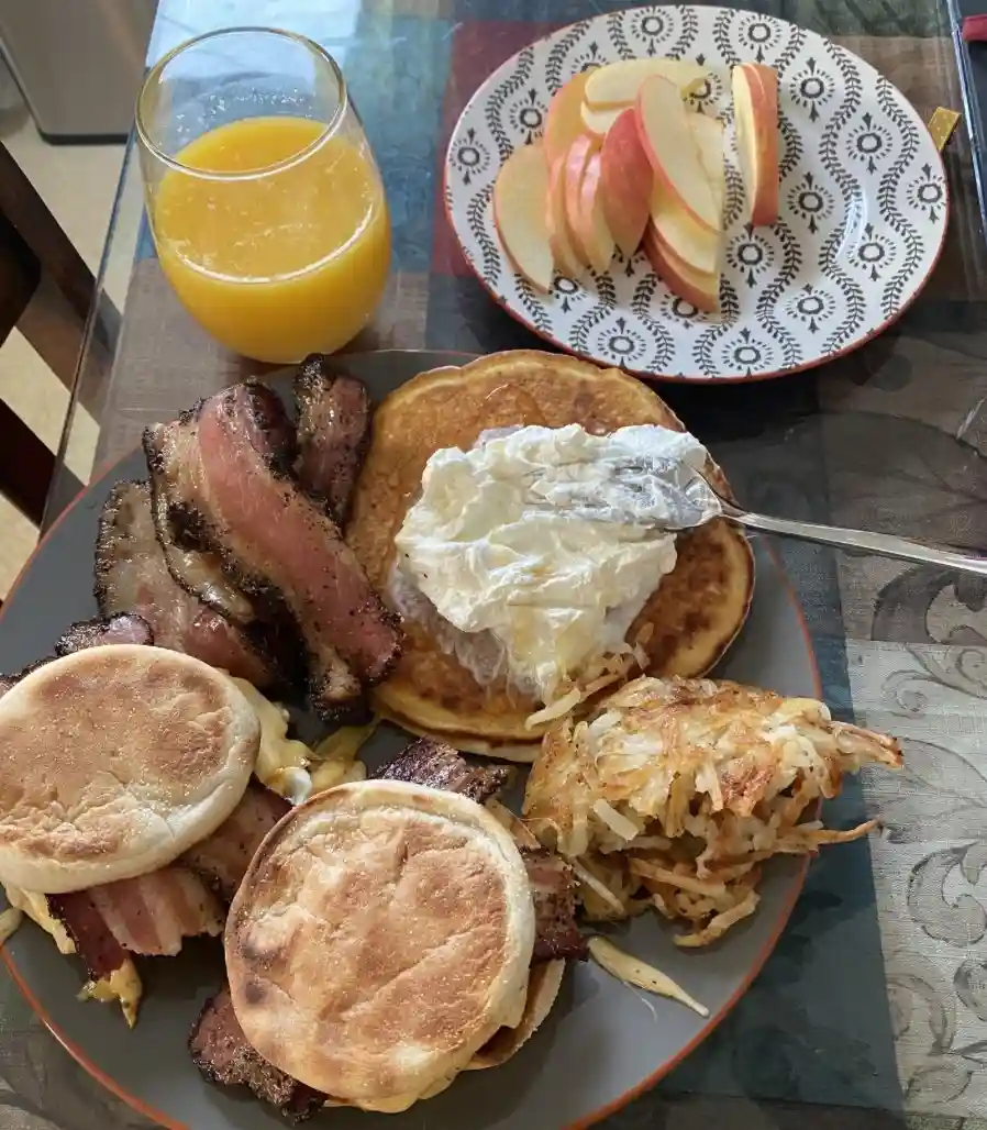 A plate of breakfast food including pancakes with whipped cream, bacon, hash browns, and English muffins, next to a glass of…