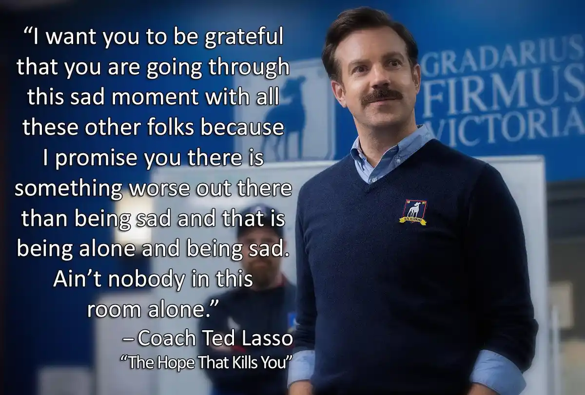 A man with a mustache wearing a blue sweater over a collared shirt stands in front of a blue wall with Latin text. Overlaid…