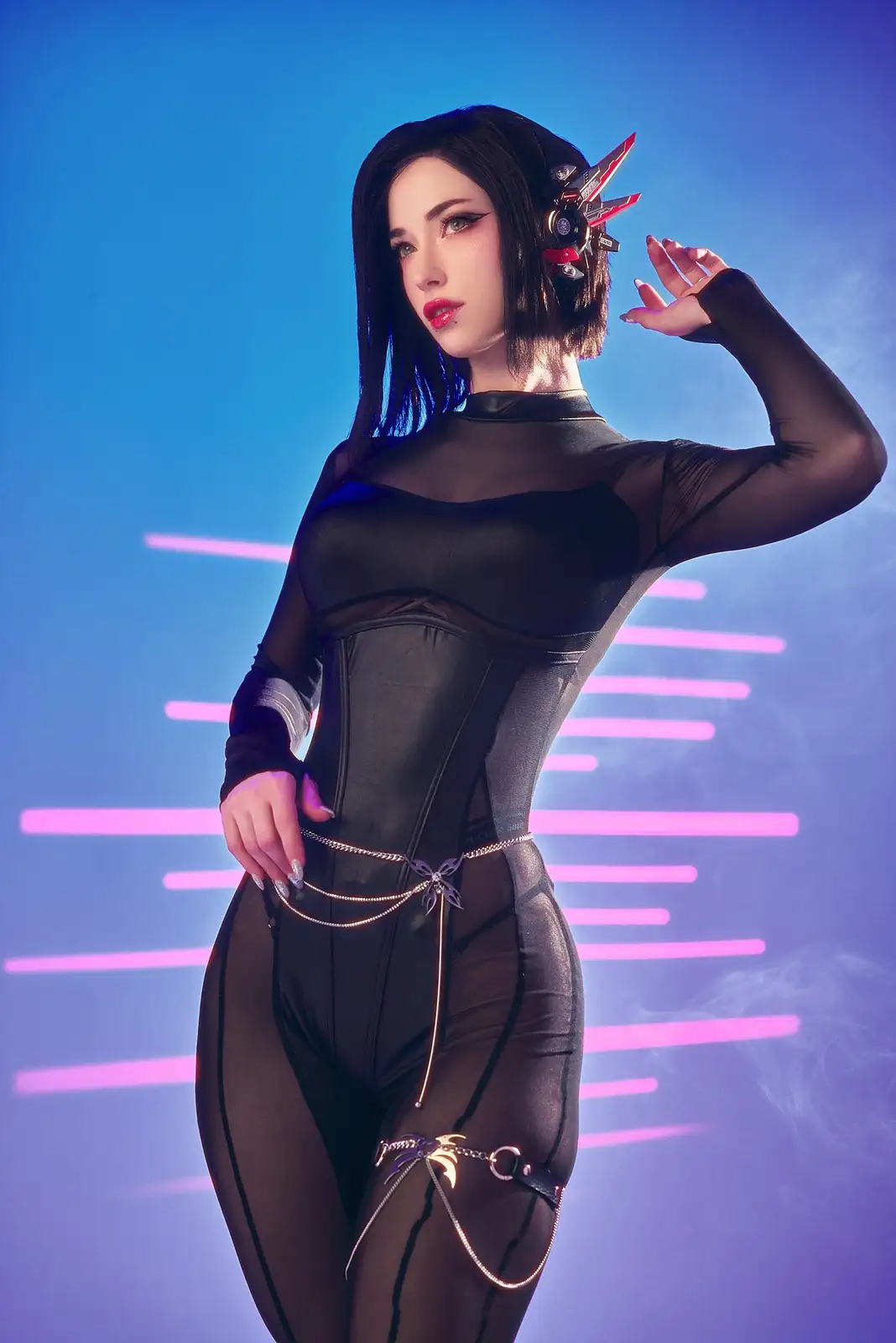 A person in a black, form-fitting, sheer bodysuit with metallic chains and a red and black mechanical headpiece poses…