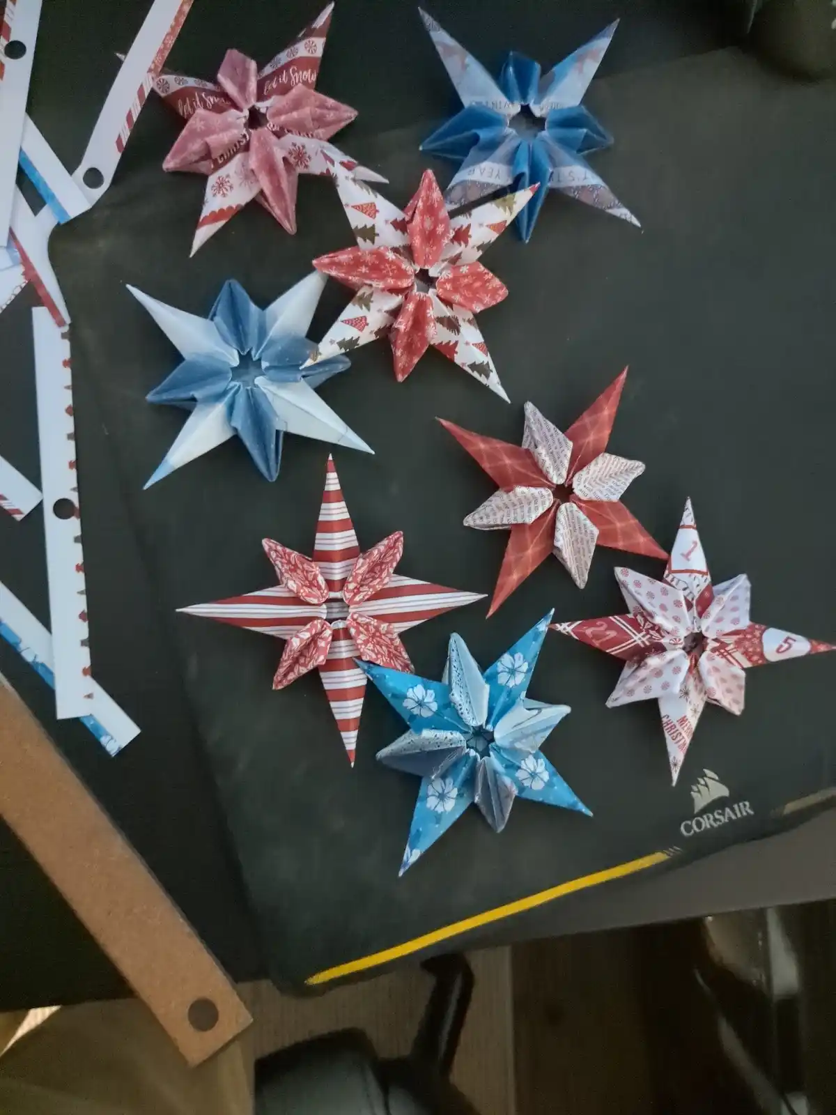 Several handmade origami stars in red, blue, and white Christmas-themed paper are arranged on a black Corsair-branded…