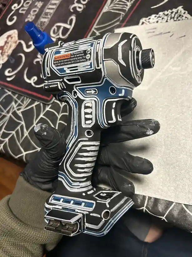 A hand in a black glove holds a power tool that has been painted to resemble a fictional character's armor, with white and…