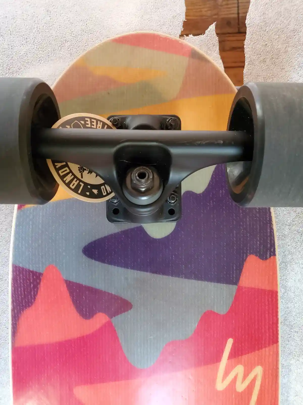 A close-up photo of a skateboard's trucks and wheels, showing a circular sticker on the truck that reads 'NO LANDY TREE' in…
