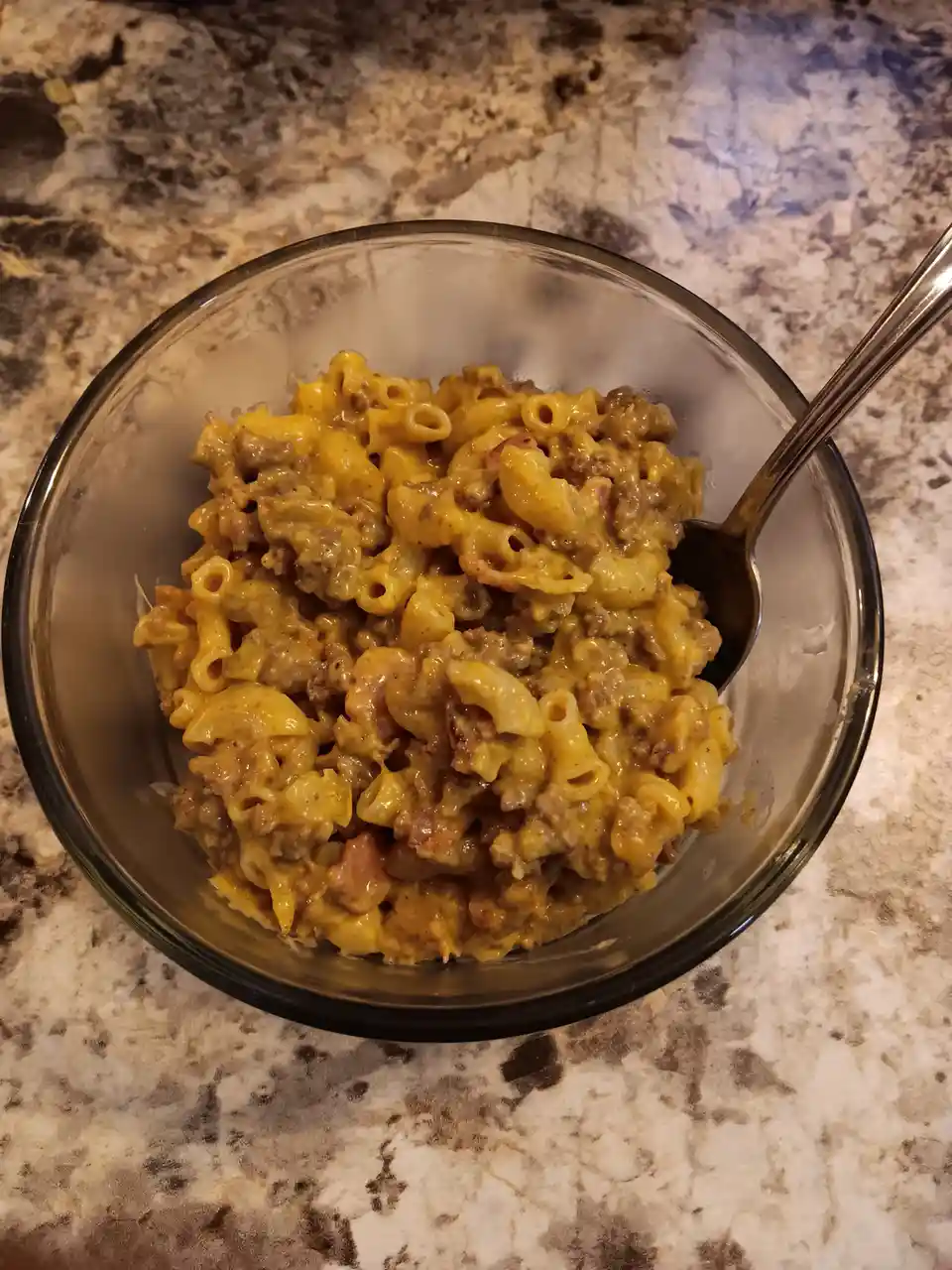 A close-up photo shows a glass bowl filled with macaroni and cheese mixed with ground meat and possibly ham, resting on a…