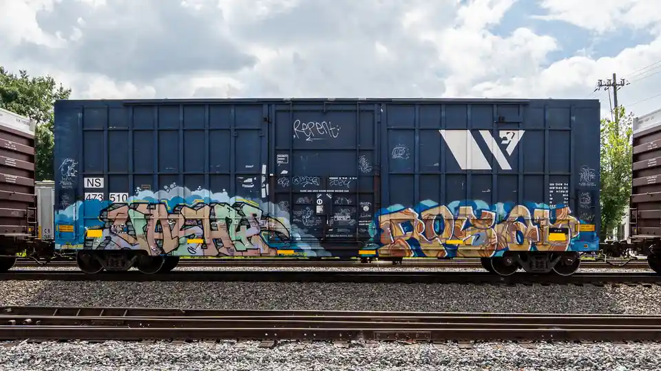 A blue freight train car covered in colorful graffiti sits on railway tracks under a cloudy sky. The word 'repent' is…