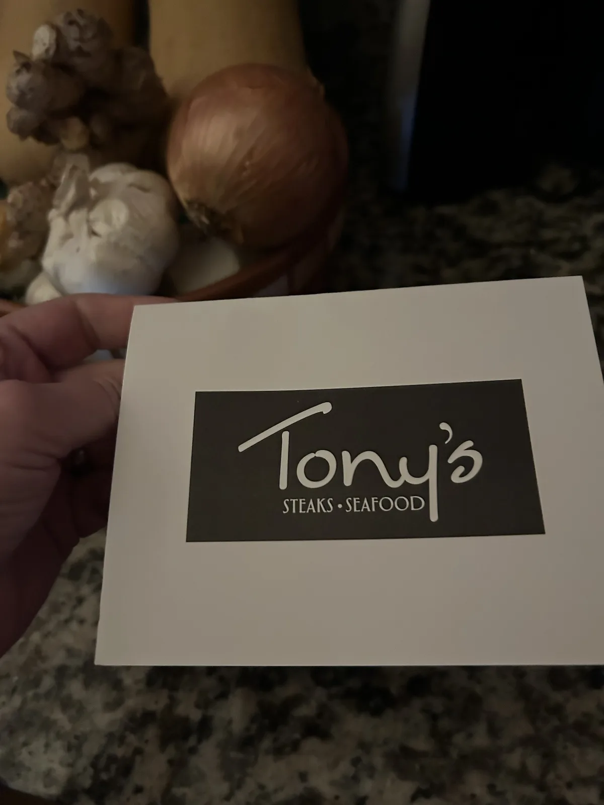 A hand holds a white card displaying a black logo that reads 'Tony's STEAKS • SEAFOOD' in white cursive and sans-serif…