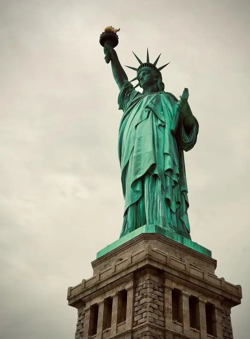 A low-angle shot of the Statue of Liberty against a cloudy sky, showing her green patina, raised torch, and spiked crown…