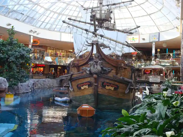A large, ornate replica of the Santa Maria ship is docked in a water feature inside a multi-level shopping mall with glass…