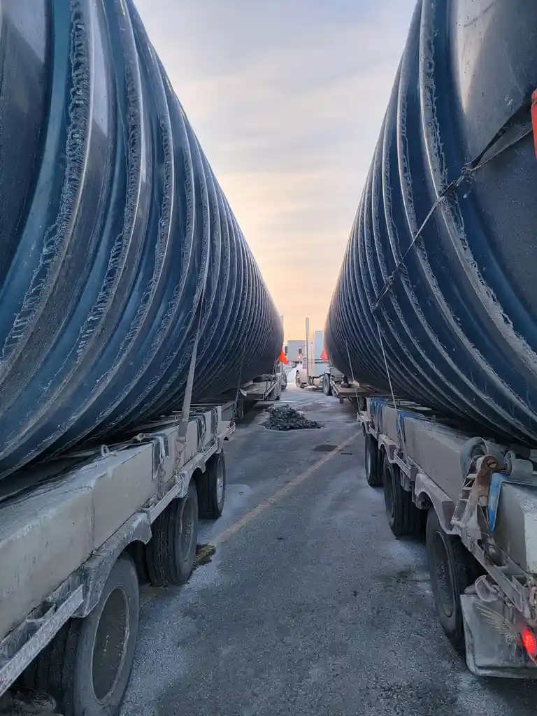 Two massive, ribbed, dark blue pipes are loaded onto flatbed trailers and parked parallel to each other, creating a narrow…