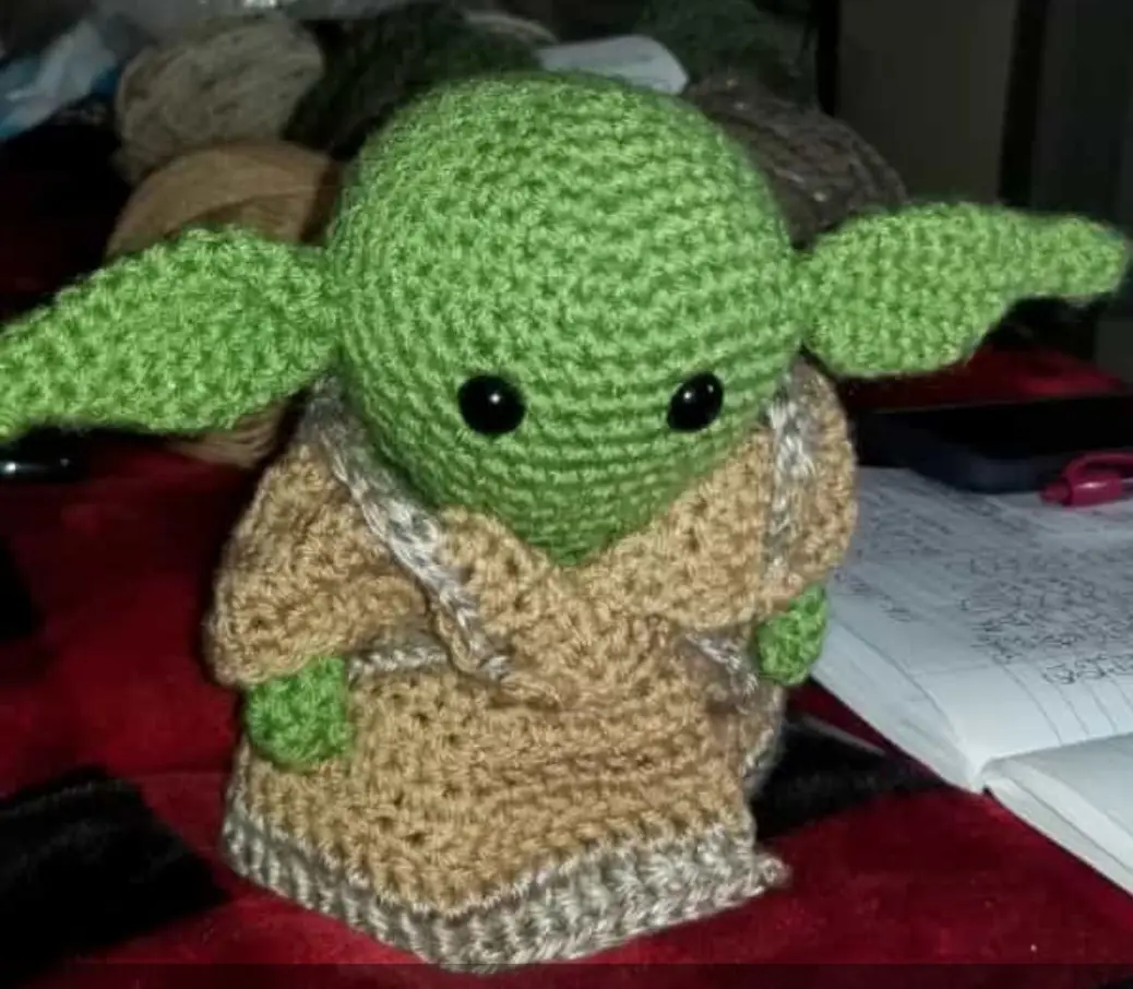 A handmade crocheted plush toy resembling the character Grogu (Baby Yoda) from Star Wars, featuring large green ears, black…