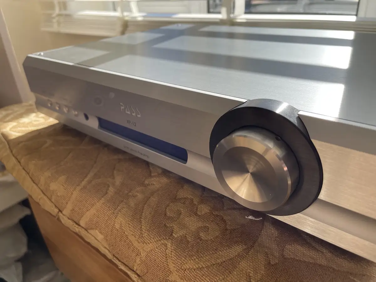 A silver Pass XP-12 Line Level Preamplifier sits on a patterned fabric surface, its large volume knob prominently featured…