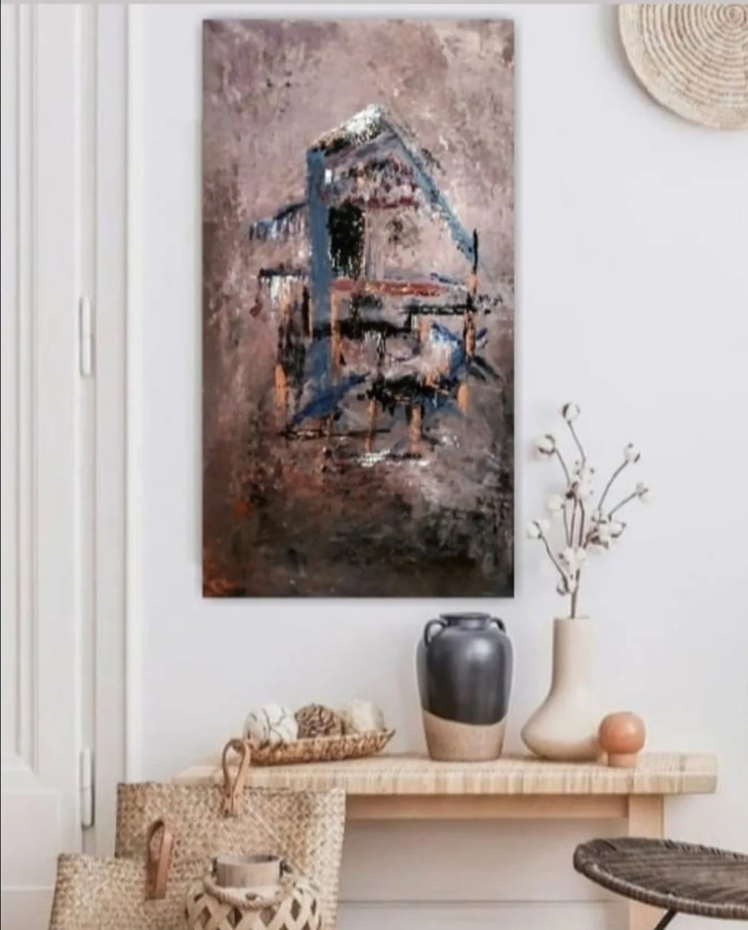 An abstract painting of a house hangs on a white wall above a wooden console table with decorative vases and baskets. The…