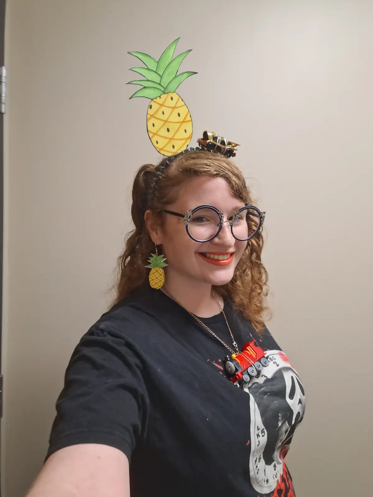 A smiling woman with curly hair and glasses wears a pineapple-shaped headband with a tiny train on top, pineapple earrings…
