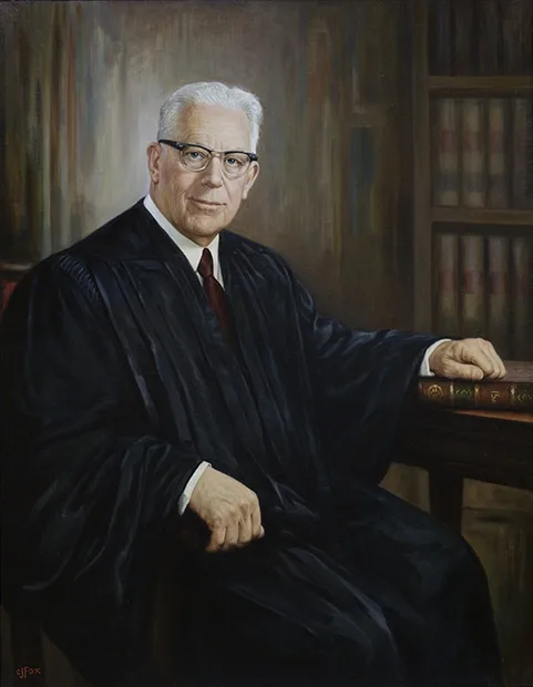 a formal portrait of an older white man with glasses, wearing a black judicial robe over a white shirt and red tie, seated…