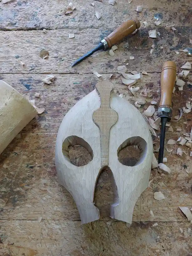 A wooden carving of a skull with eye sockets and a mouth, sitting on a workbench covered in wood shavings, with two chisels…