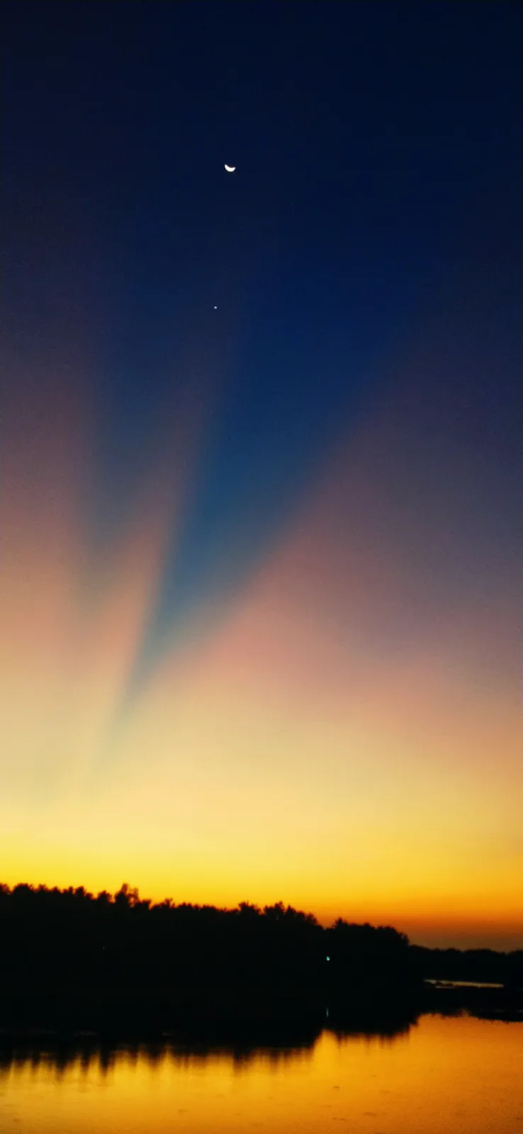 A serene twilight sky with a crescent moon and a bright star above a silhouetted treeline reflected in calm water, overlaid…