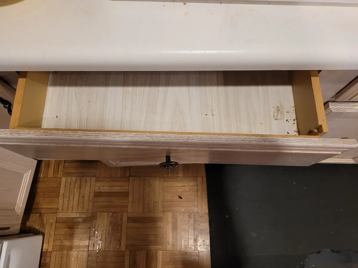 A close-up photo shows an empty, slightly dirty wooden drawer pulled open from a white cabinet, revealing its bare bottom…