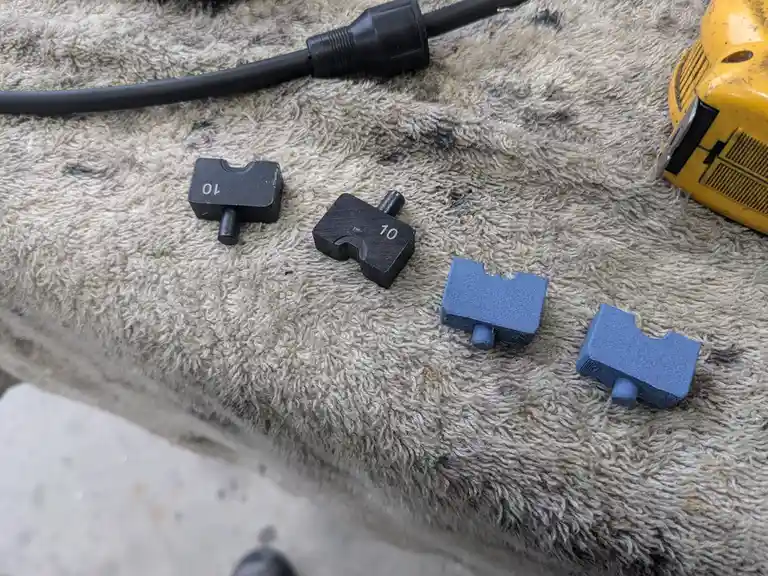 Four small, blocky mechanical parts—two black and two blue—are arranged on a worn, fluffy beige towel. The black parts are…