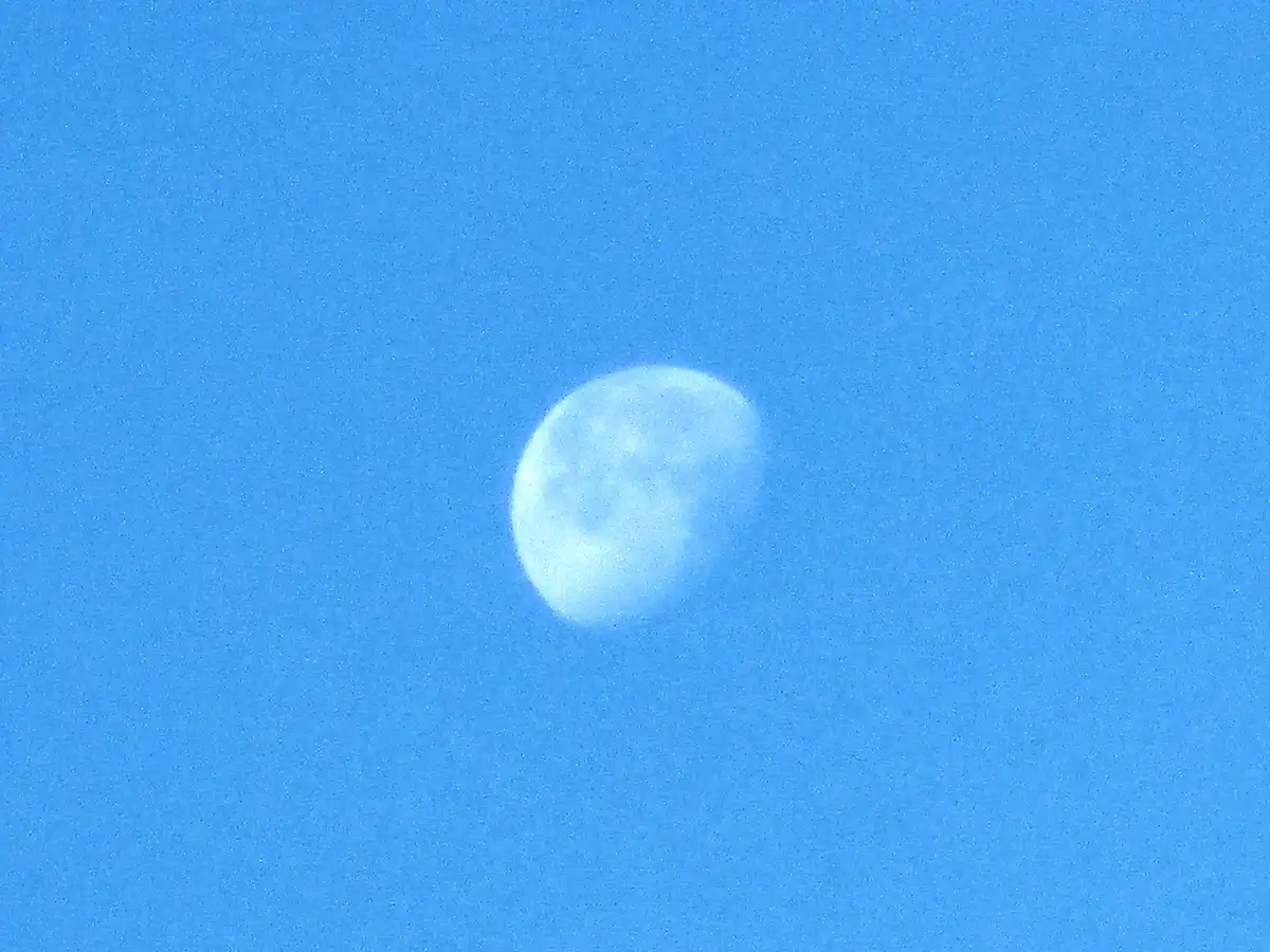 A pale, slightly hazy moon is centered in a bright blue sky, which has a grainy texture. The overlaid text reads 'the moon…