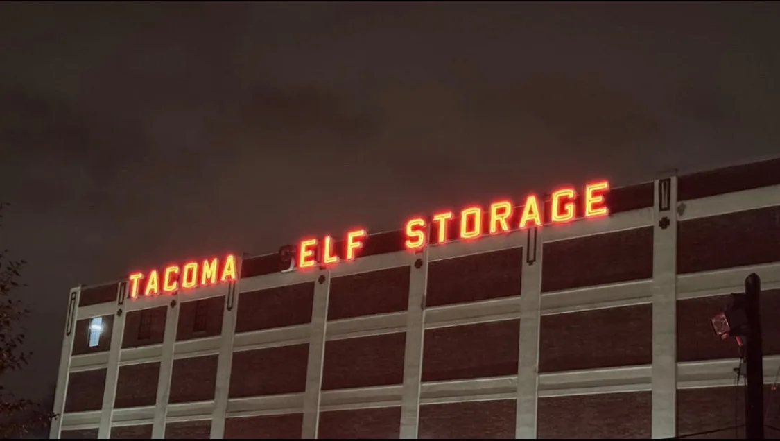 A nighttime photo of a brick building with a bright orange neon sign that reads 'TACOMA ELF STORAGE', where the word 'SELF'…