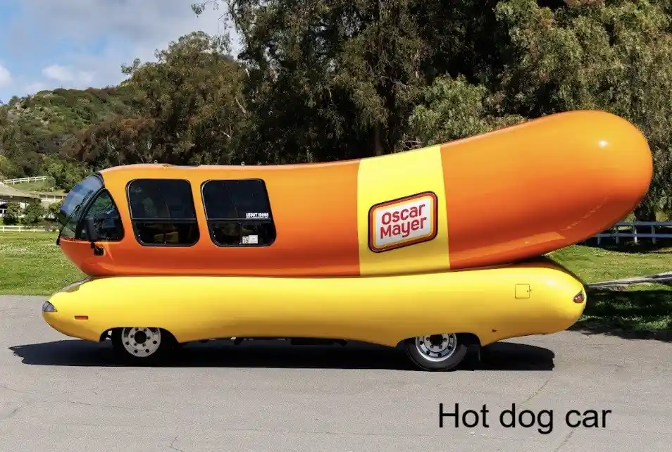 A custom vehicle shaped like a hot dog in a bun, painted orange and yellow with an 'Oscar Mayer' logo on the side, parked…