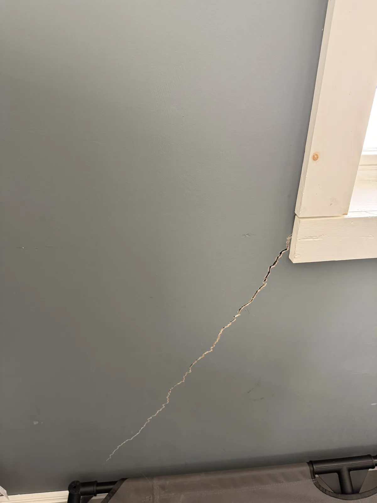 A close-up photo shows a long, jagged crack running diagonally across a gray-painted interior wall, starting near the bottom…